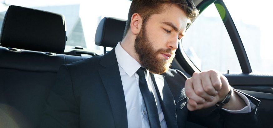Confident young businessman looking at his watch while sitting on the back seat of a car